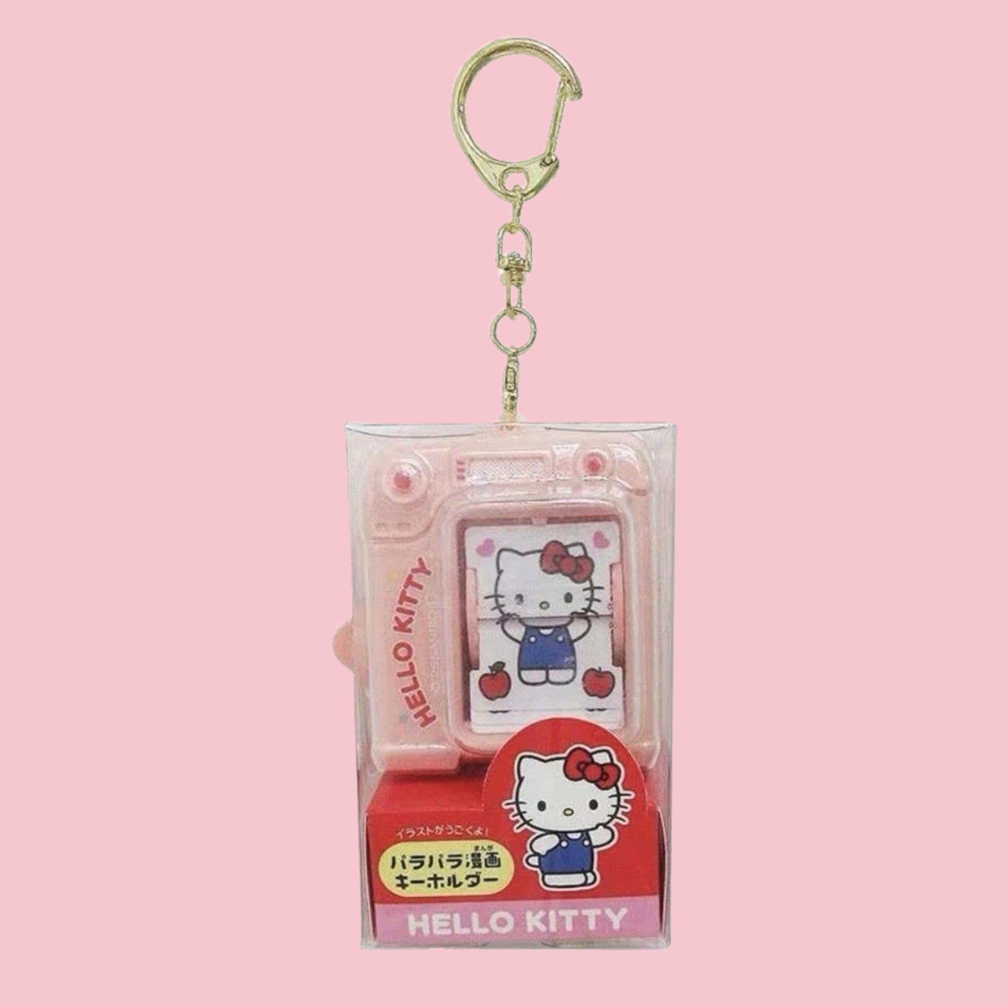 Pink plastic camera shaped keychain in original packaging, showing Hello Kitty in blue overalls flipping through a small illustrated booklet inside the window, with a red Hello Kitty character card and Japanese text on the box, against a solid pink background.
