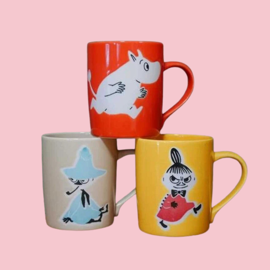 Three Japanese Moomin mugs stacked together showing Moomin on a red mug, Snufkin on a grey mug, and Little My on a yellow mug, on a pink background