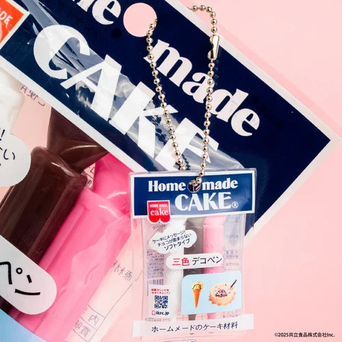 A miniature toy cake decoration kit labeled 'Home made CAKE' with a silver ball chain, shown alongside the full size cake decoration kit on a light pink background.