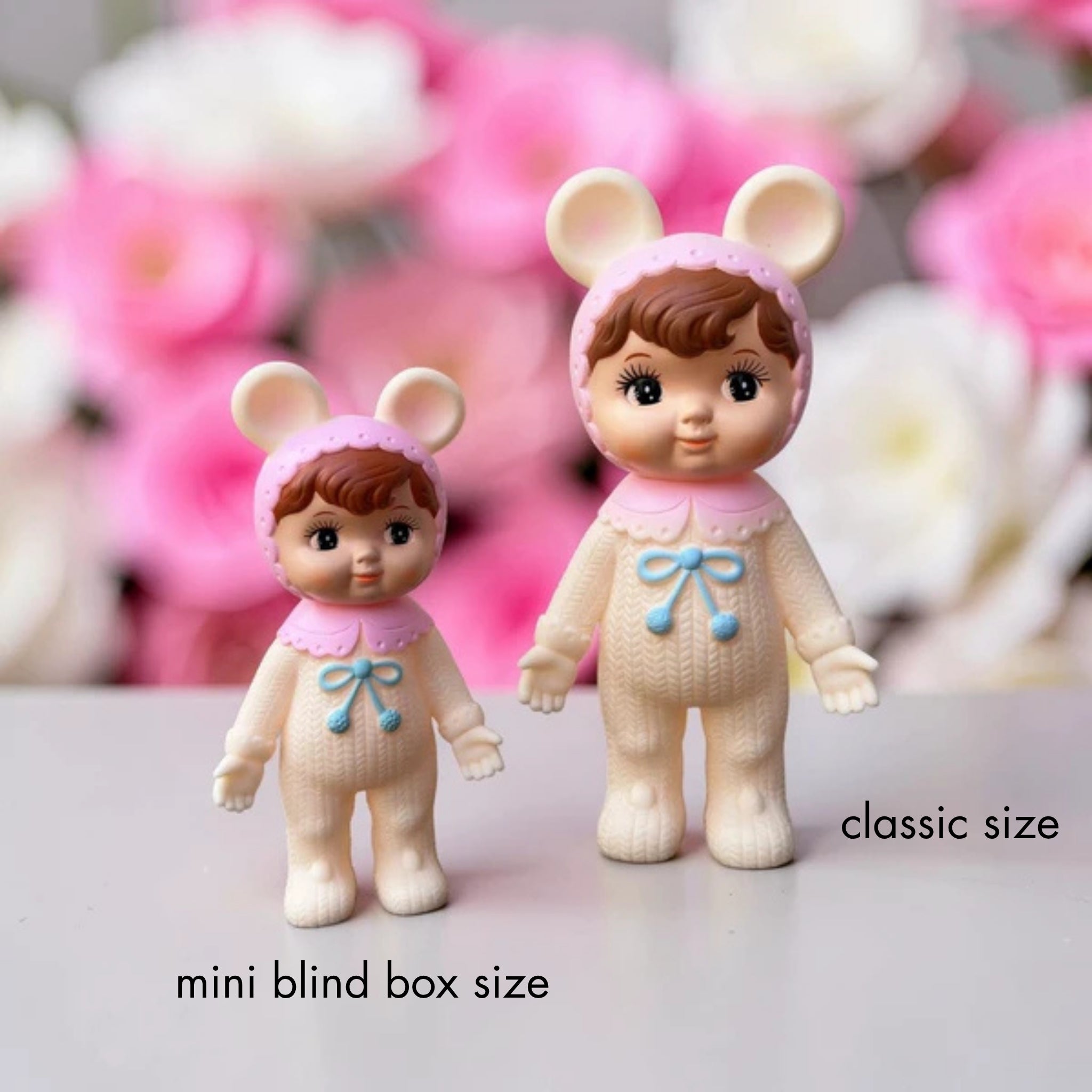 Two Nakayoshi Charmy dolls in matching pink bunny-ear hoods and cream knit outfits with blue bow details, shown side by side against a soft floral background to compare sizes, labeled "mini blind box size" for the smaller doll and "classic size" for the larger one.