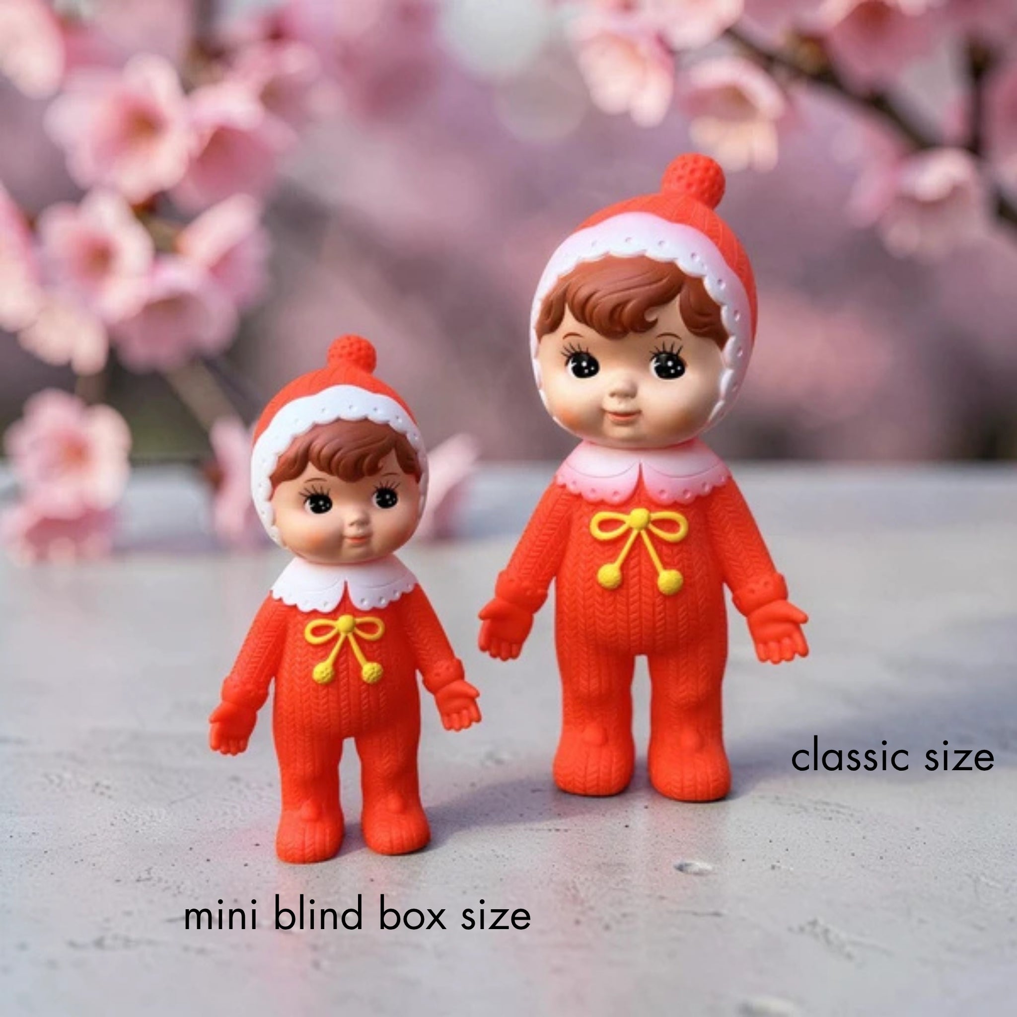 Two Nakayoshi Charmy dolls in matching red pom-pom hoods and knit outfits with yellow bow details, standing side by side against a cherry blossom background to show the size difference, labeled "mini blind box size" for the smaller doll and "classic size" for the larger one.