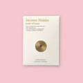 Kousaido brass incense holder on its white product card, showing the disc-shaped holder and text reading "Incense Holder made of brass," with "Kousaido Kyoto Japan" printed at the bottom, against a pink background