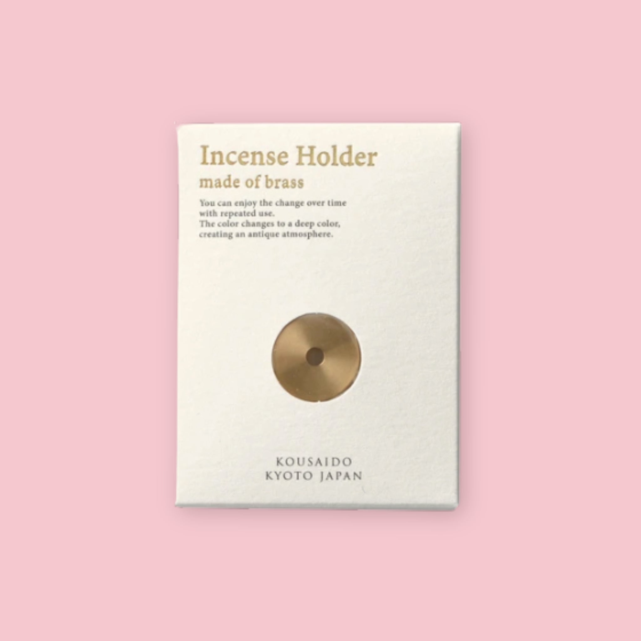 Kousaido brass incense holder on its white product card, showing the disc-shaped holder and text reading "Incense Holder made of brass," with "Kousaido Kyoto Japan" printed at the bottom, against a pink background