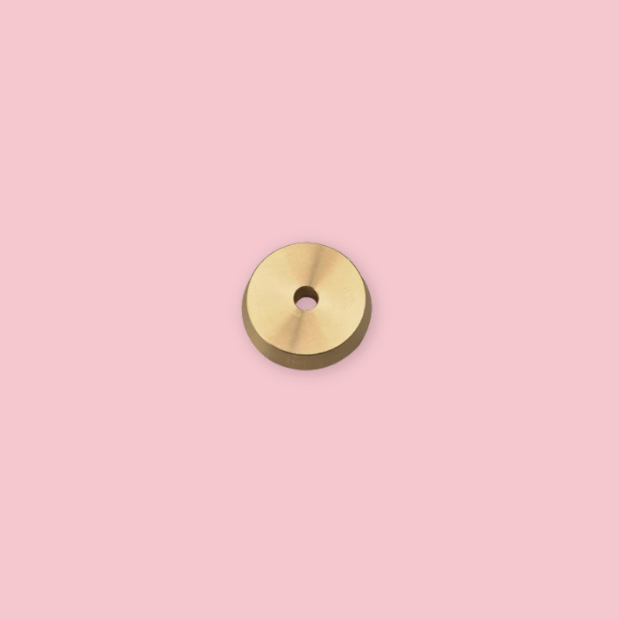 Small circular brass incense holder disc with a center hole for stick incense, shown against a pink background, with warm gold tones