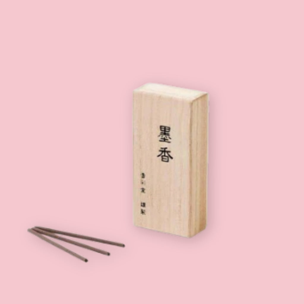 Kousaido Calligraphy Ink incense in a light paulownia wood box printed with the kanji 墨香, with three dark charcoal-brown incense sticks fanned out in front, against a pink background
