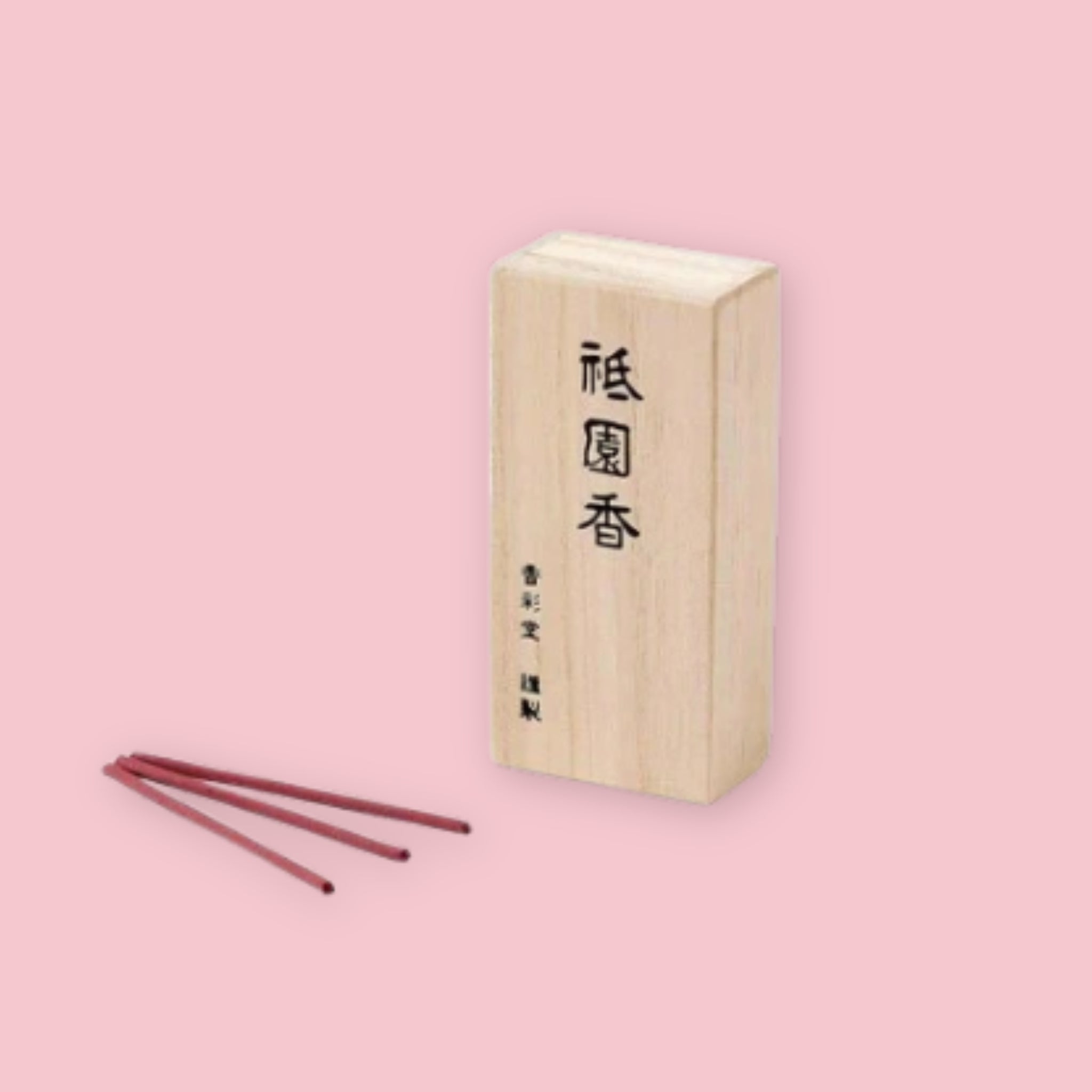 Kousaido Gion Night incense in a light paulownia wood box printed with the kanji 祇園香, with three dark reddish-brown incense sticks fanned out in front, against a pink background