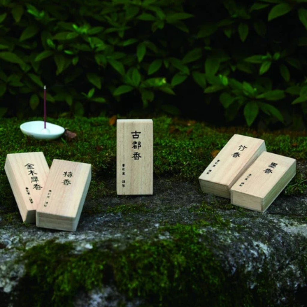 Five Kousaido incense boxes with kanji lettering arranged on a moss-covered stone outdoors, with a single lit incense stick burning in a small dish to the left, green foliage in the background.