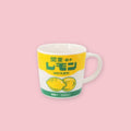 A white ceramic mug featuring a retro Japanese label design with yellow and green colorblocking, bold green text reading 関東・栃木 レモン (Kanto Tochigi Lemon), illustrated lemons, and 500ml lemon milk drink branding, displayed on a pink background.