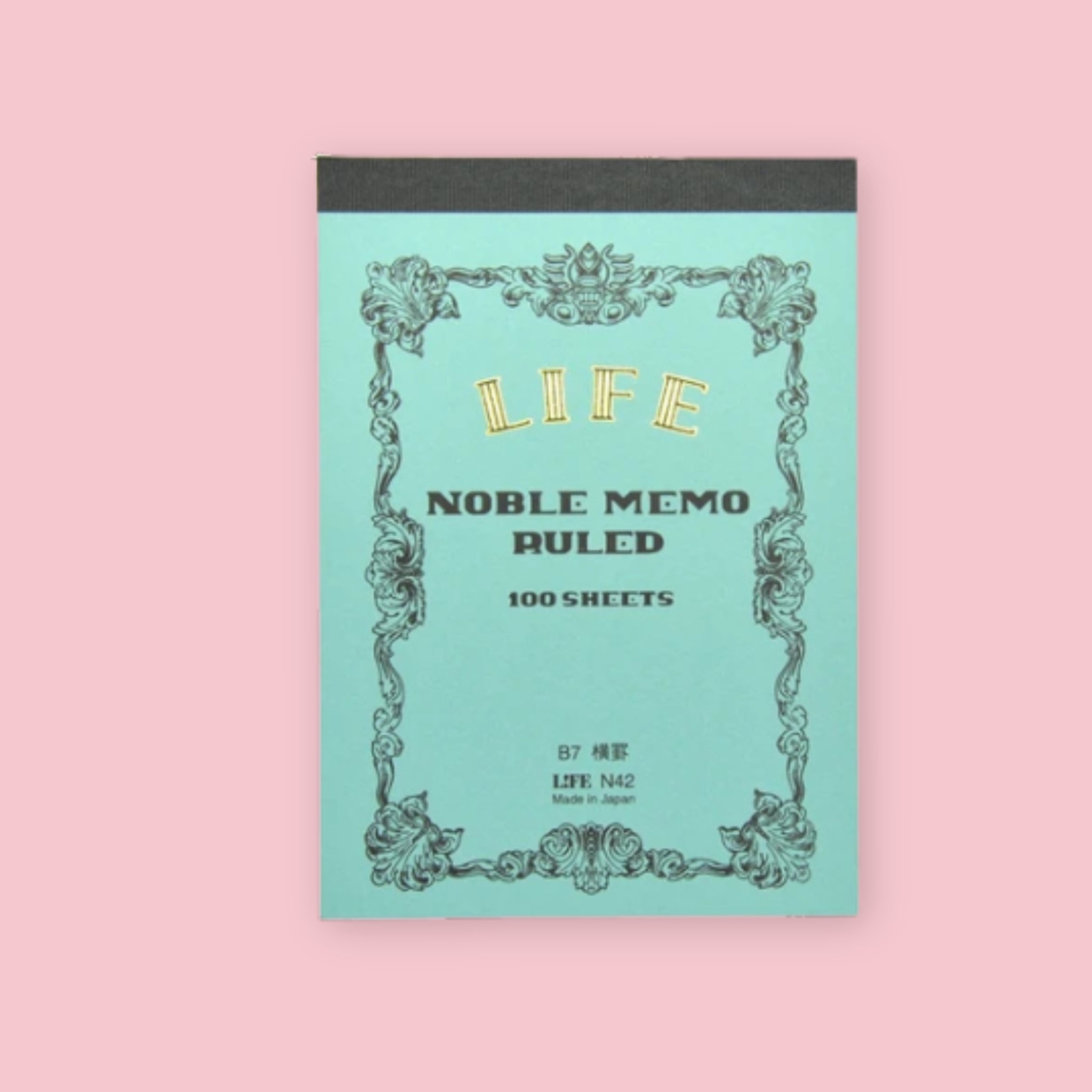 Light blue LIFE Noble Memo ruled notepad with ornate vintage-style border and black top binding on a pale pink background.