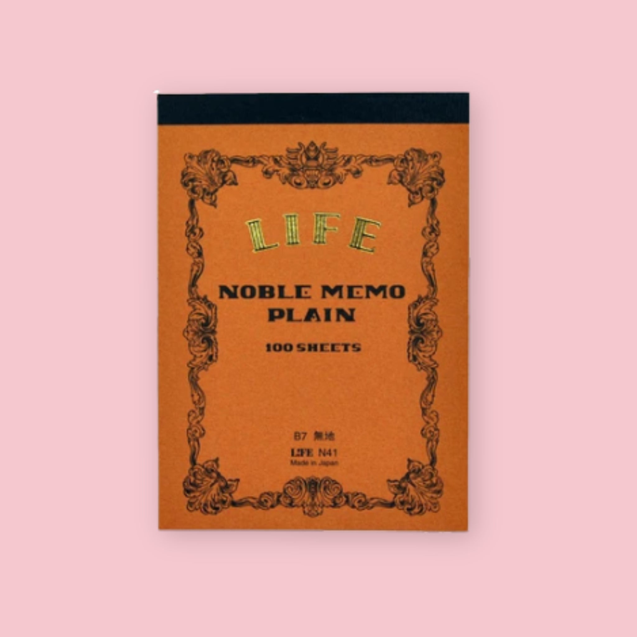 Brown LIFE Noble Memo plain notepad with ornate vintage-style border and dark top binding on a pale pink background.