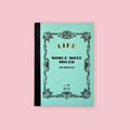 Light blue LIFE Noble Note ruled notebook with ornate vintage-style black border and black cloth spine on a pale pink background.