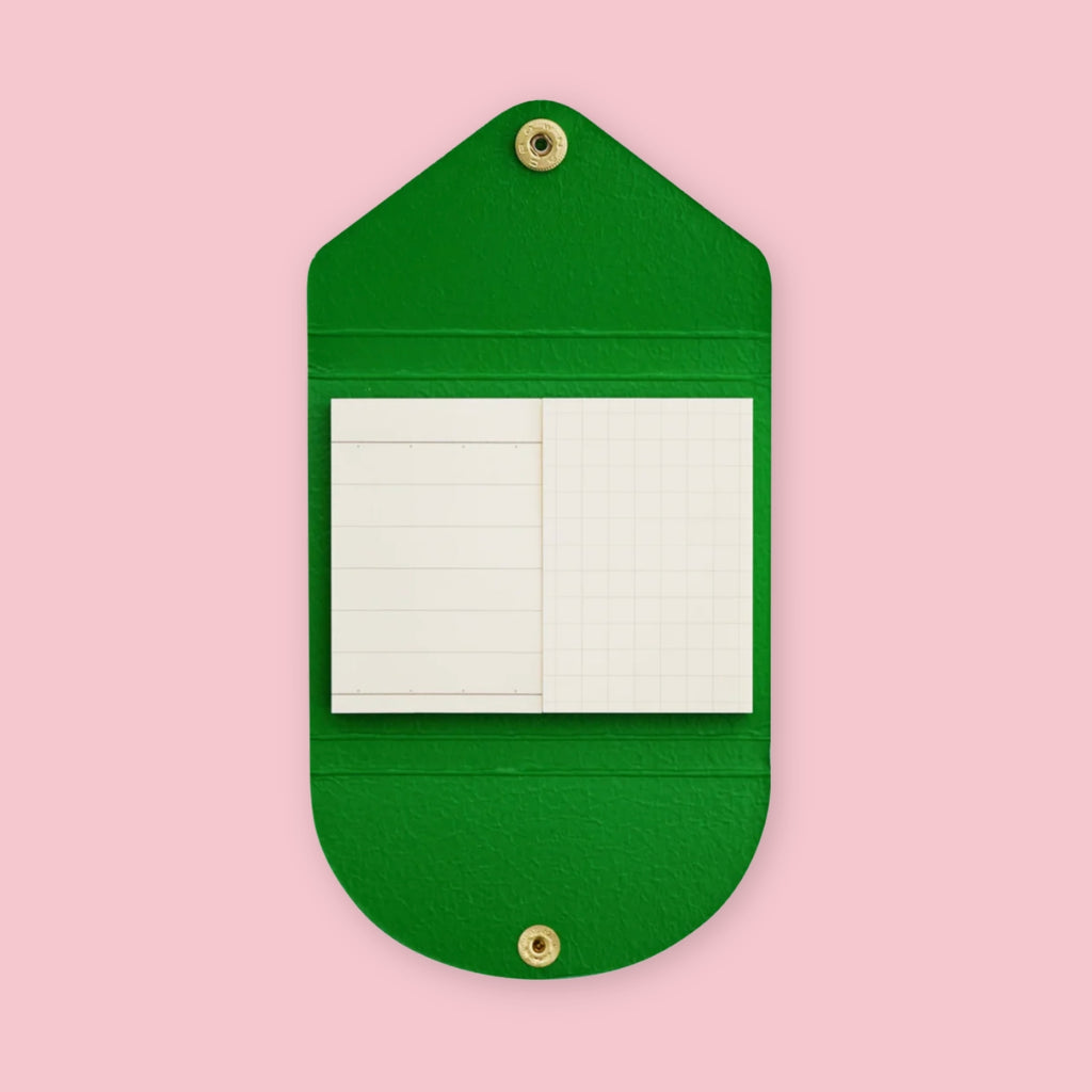 Green faux leather sticky note pouch shaped like an envelope, open to show lined and grid notepads inside, snap closures top and bottom, on a pink background.
