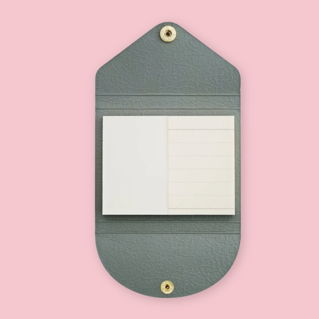 Grey faux leather sticky note pouch shaped like an envelope, open to show plain and lined notepads inside, snap closures top and bottom, on a pink background.