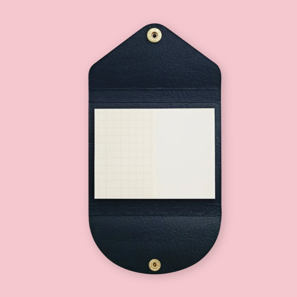 Navy faux leather sticky note pouch shaped like an envelope, open to show grid and plain notepads inside, snap closures top and bottom, on a pink background.