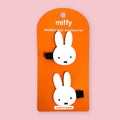 Two white bunny hair clips with black elastic bands, mounted on an orange card labeled 'miffy BANGS Hair accessories' against a light pink background.