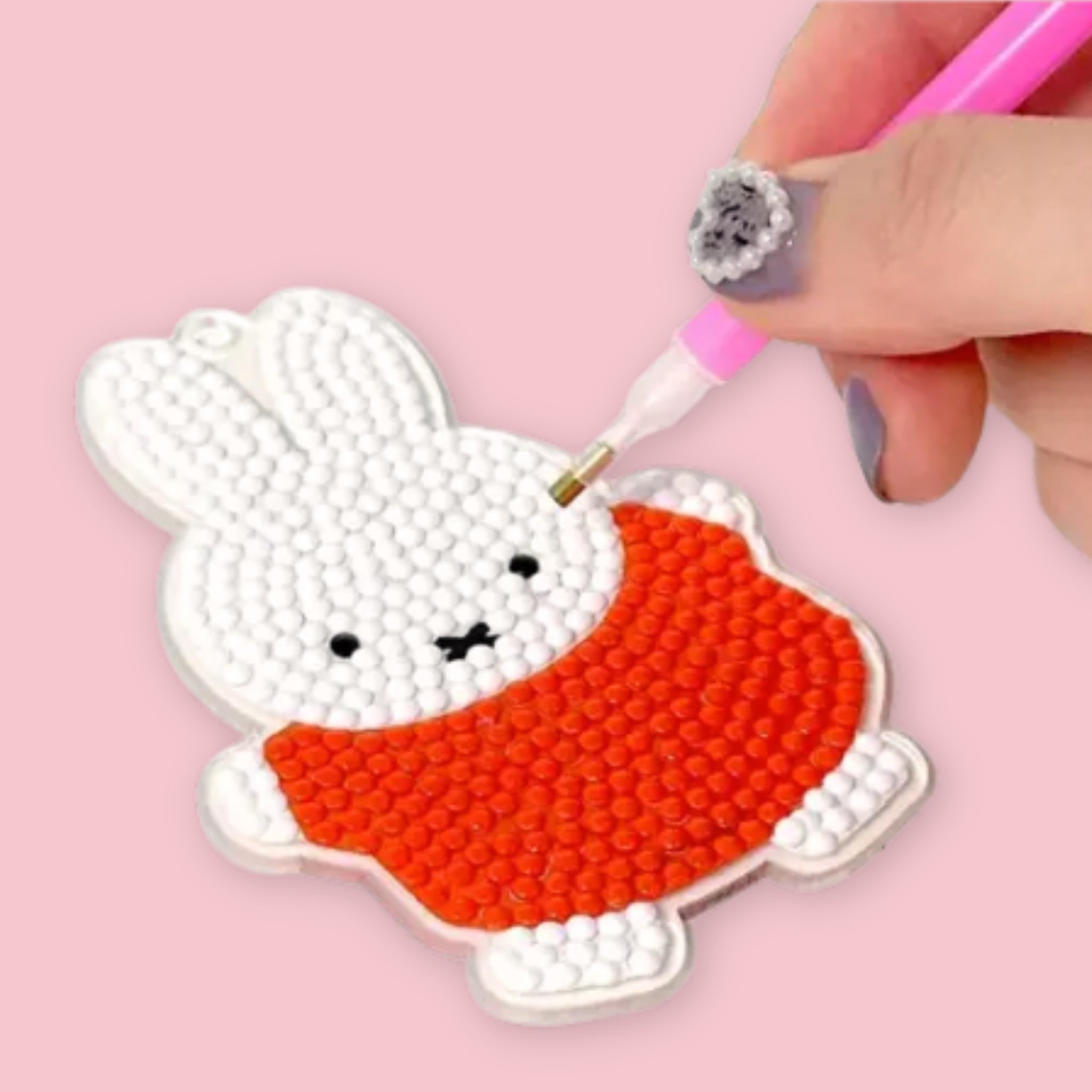 A hand using a pink applicator pen to place gem beads onto a Miffy keychain acrylic base, showing the white bunny in an orange dress nearly complete, against a pink background