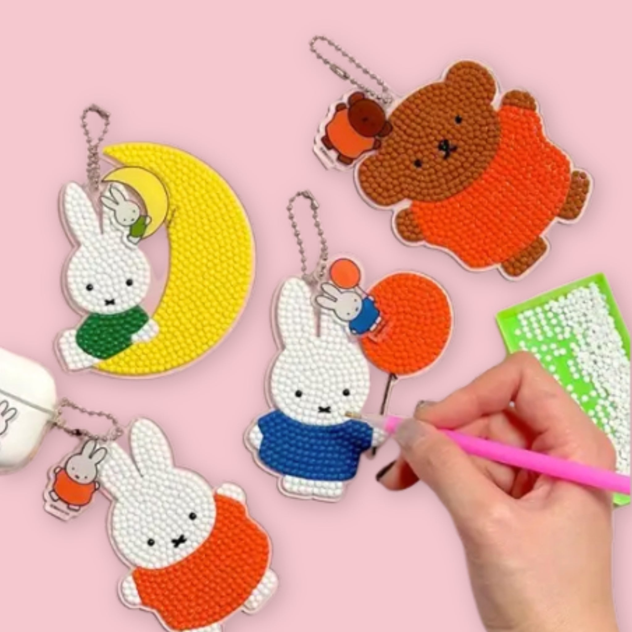 Four completed diamond painting keychains laid out on a pink background alongside a hand working on a green acrylic base, showing the Miffy on the Moon, Boris, Miffy with Balloon, and Miffy designs covered in gem beads