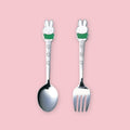 A stainless steel spoon and fork set, each with a Miffy character handle topper wearing a green outfit and floral engraving on the stem, displayed upright side by side on a pink background.