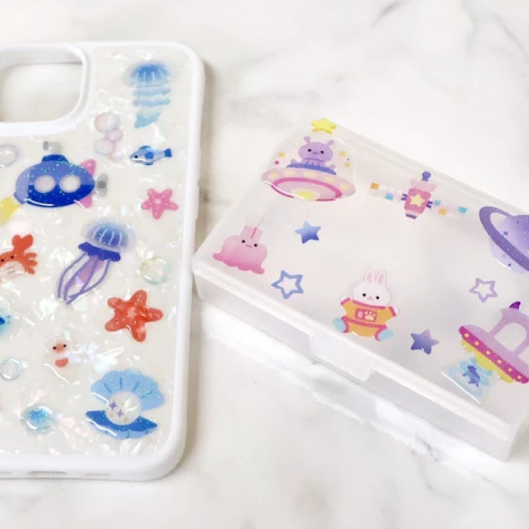 A clear phone case with colorful sea life and space-themed charms, shown next to its matching translucent storage box on a white marble surface.