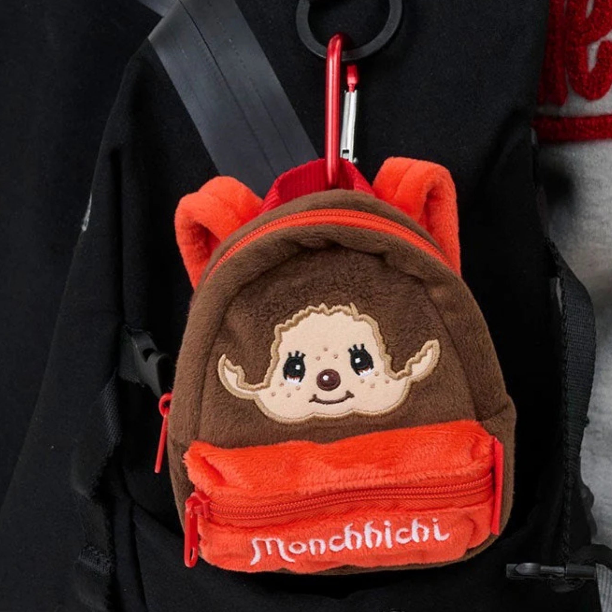 Monchhichi plush backpack pouch clipped to a black backpack, showing the embroidered face and red front zipper pocket.