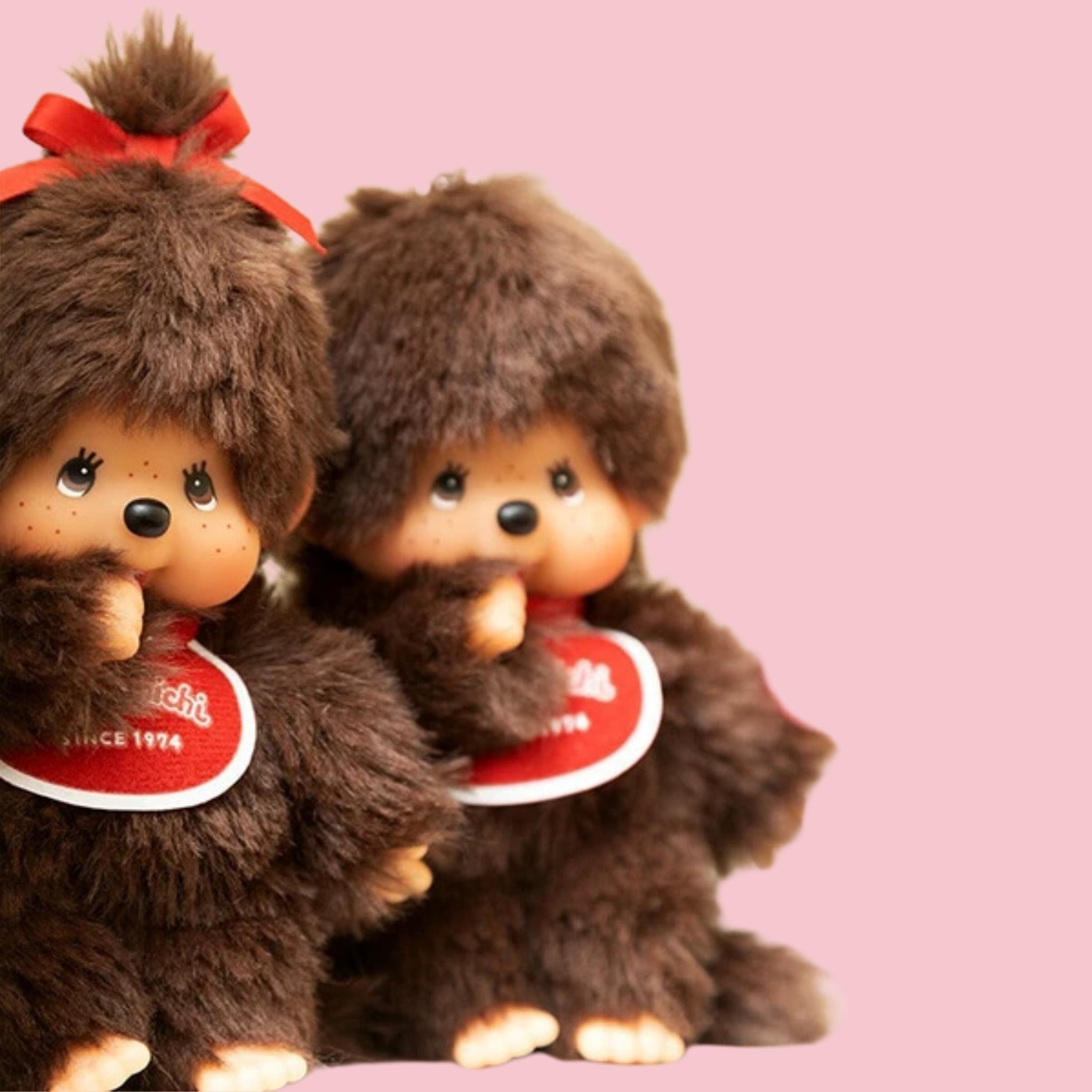 Close-up of two classic brown Monchhichi plush dolls with red bibs, shown on a pink background.