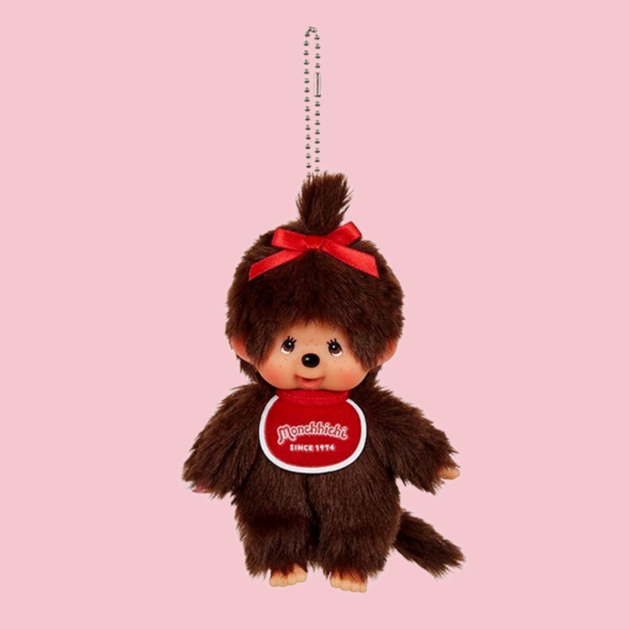 Classic brown Monchhichi girl mascot charm with red bow, red bib, and ball chain on a pink background.