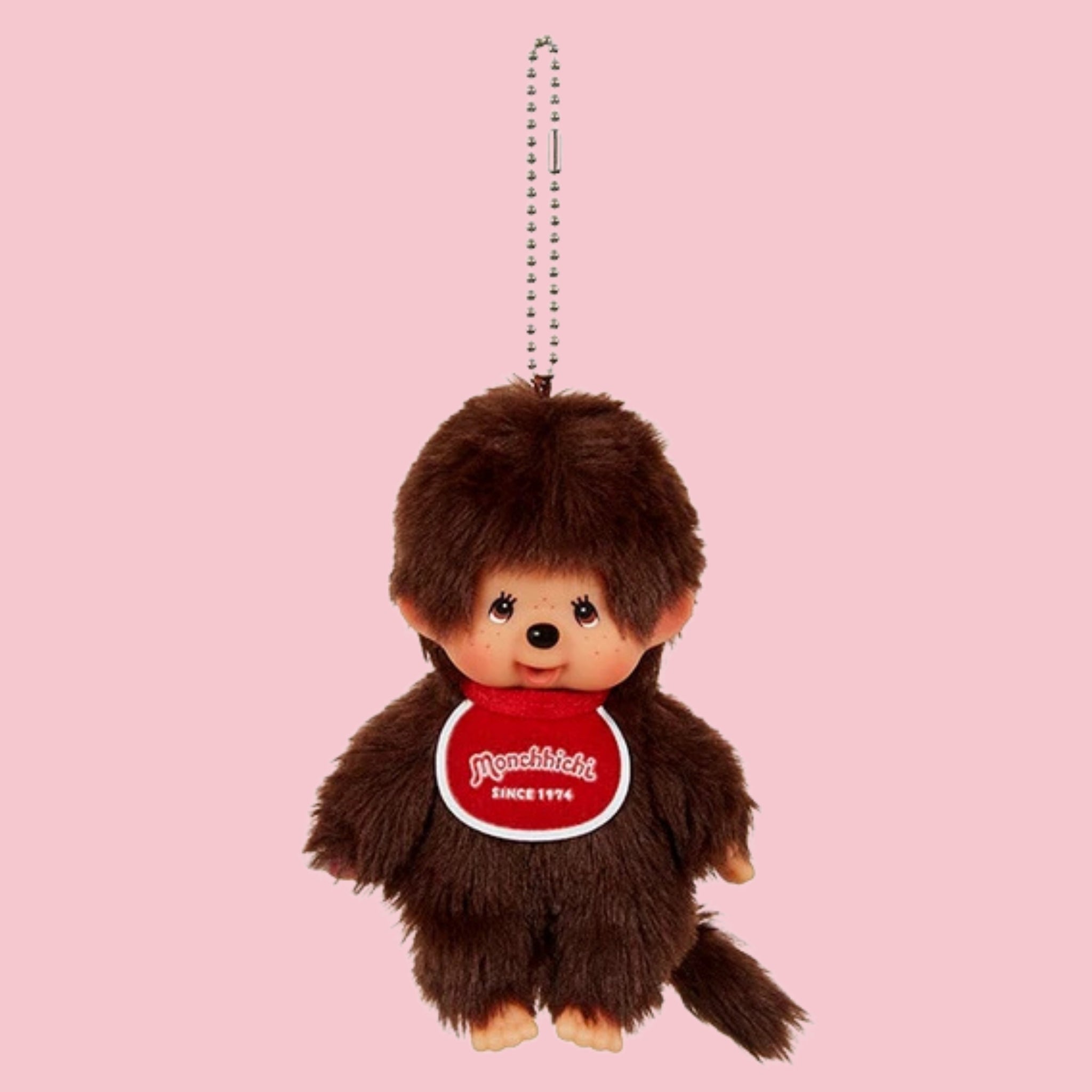 Classic brown Monchhichi boy mascot charm with red bib, ball chain, and tail on a pink background.