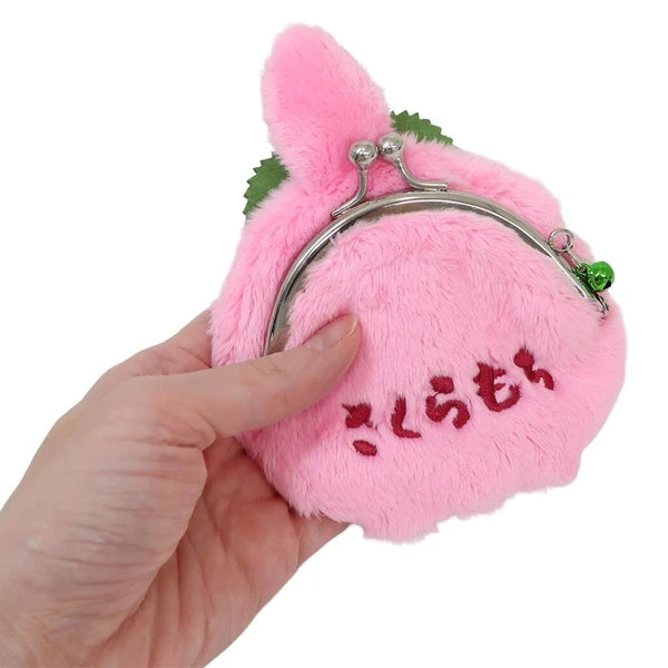 A hand holding a small pink plush coin purse shaped like a Monchhichi, with green leaf accents, a silver metal kiss-lock closure, and red embroidered Japanese text on the front, against a plain white background.