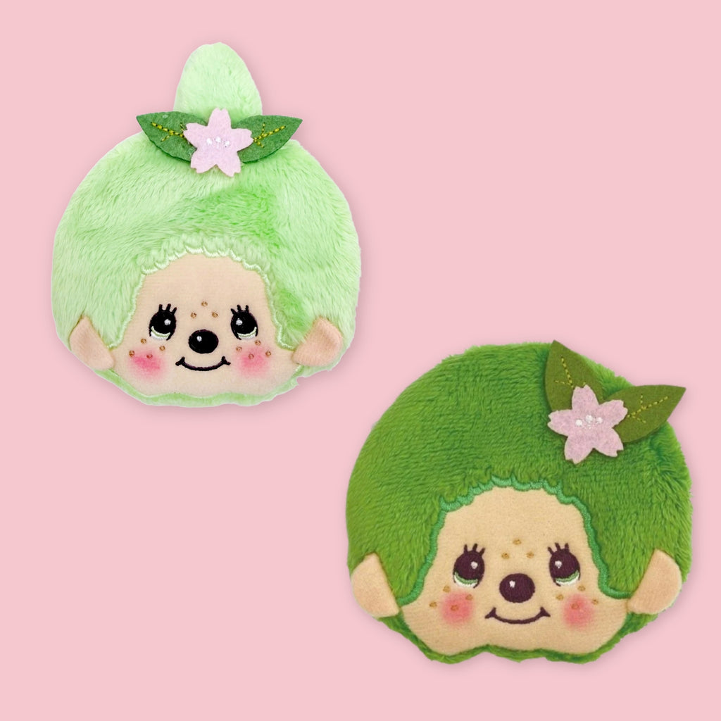 Two Monchhichi matcha coin pouches in light green and dark green, each with a pink flower and leaf hair accessory, shown on a pink background.