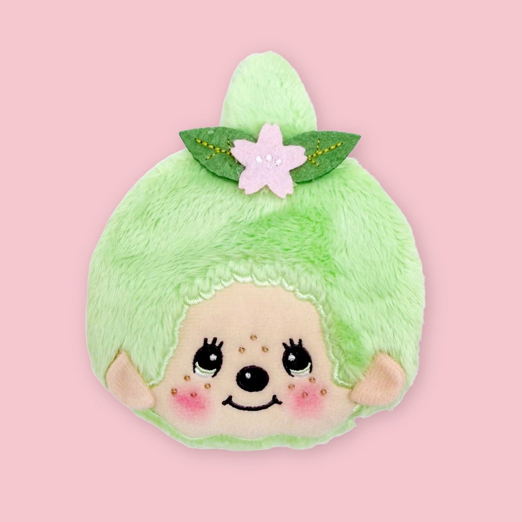 Light green Monchhichi matcha coin pouch with embroidered face and pink flower hair accessory, shown on a pink background.