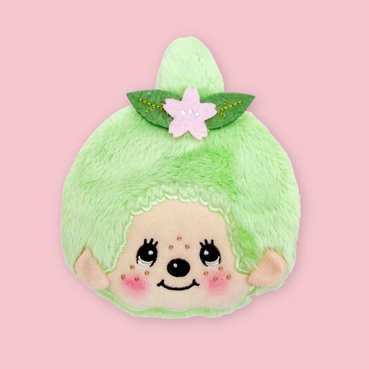 Light green Monchhichi matcha coin pouch with embroidered face and pink flower hair accessory, shown on a pink background.