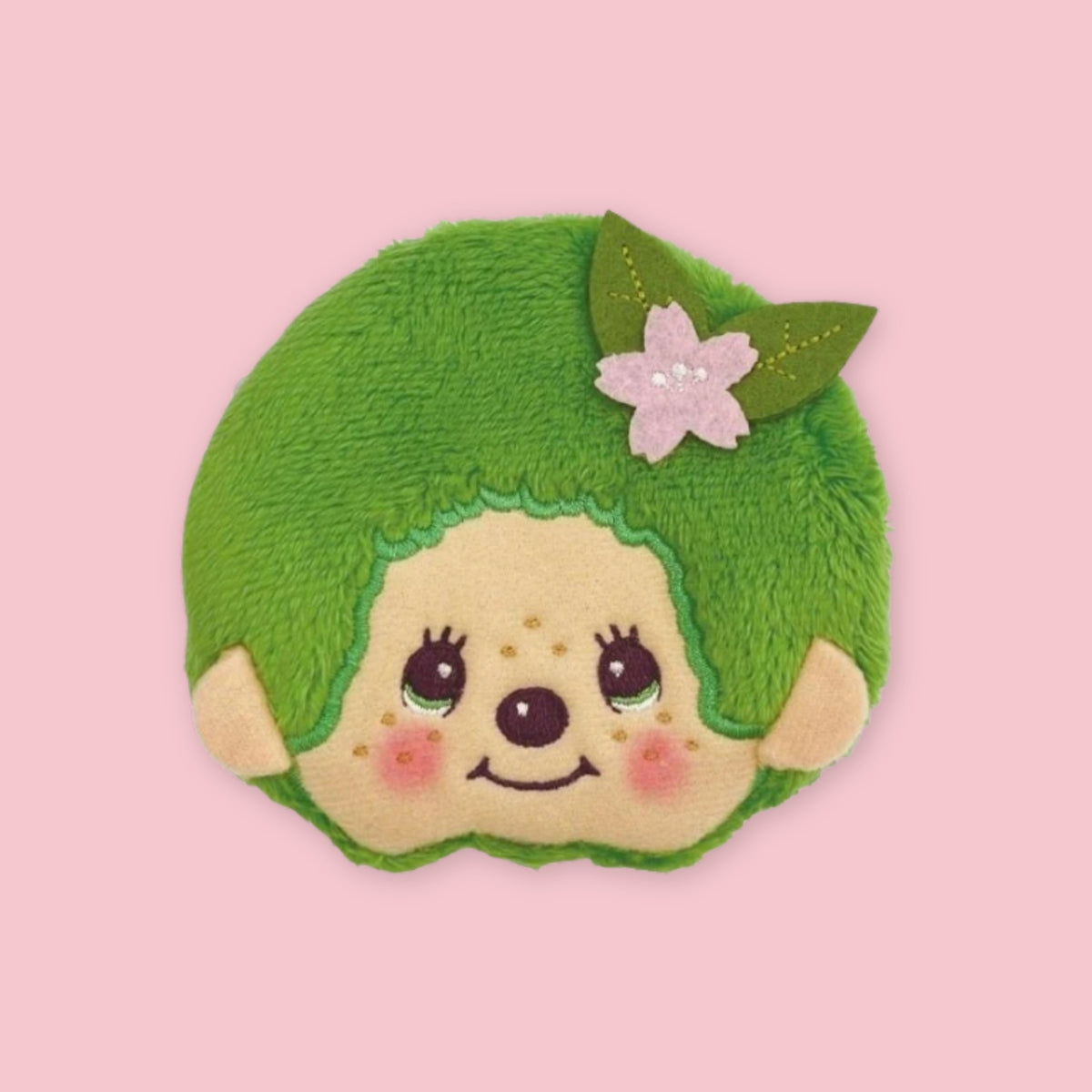 Dark green Monchhichi matcha coin pouch with embroidered face and pink flower hair accessory, shown on a pink background.