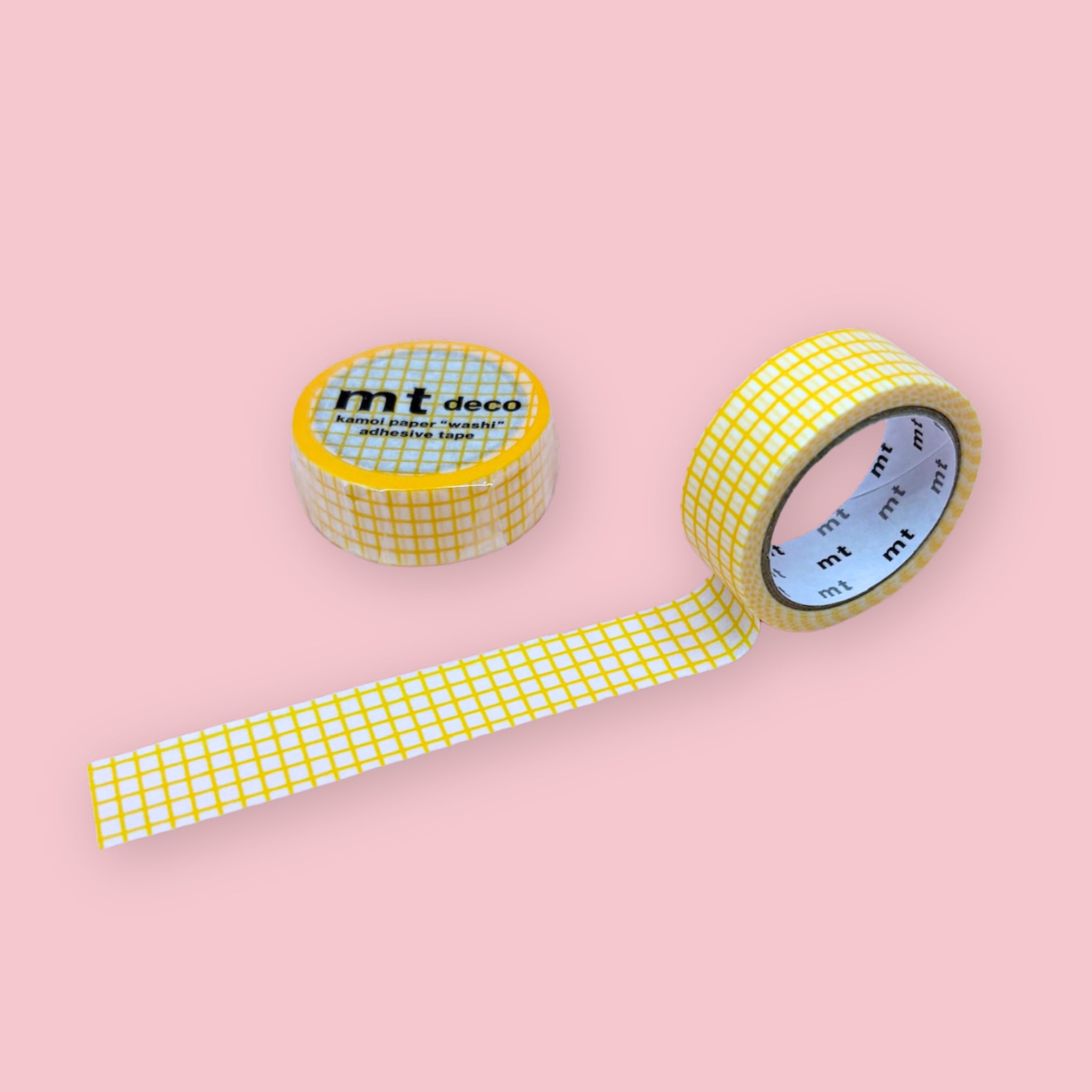 mt deco washi tape with a yellow grid pattern on white, shown partly unspooled beside a wrapped roll on a pink background.
