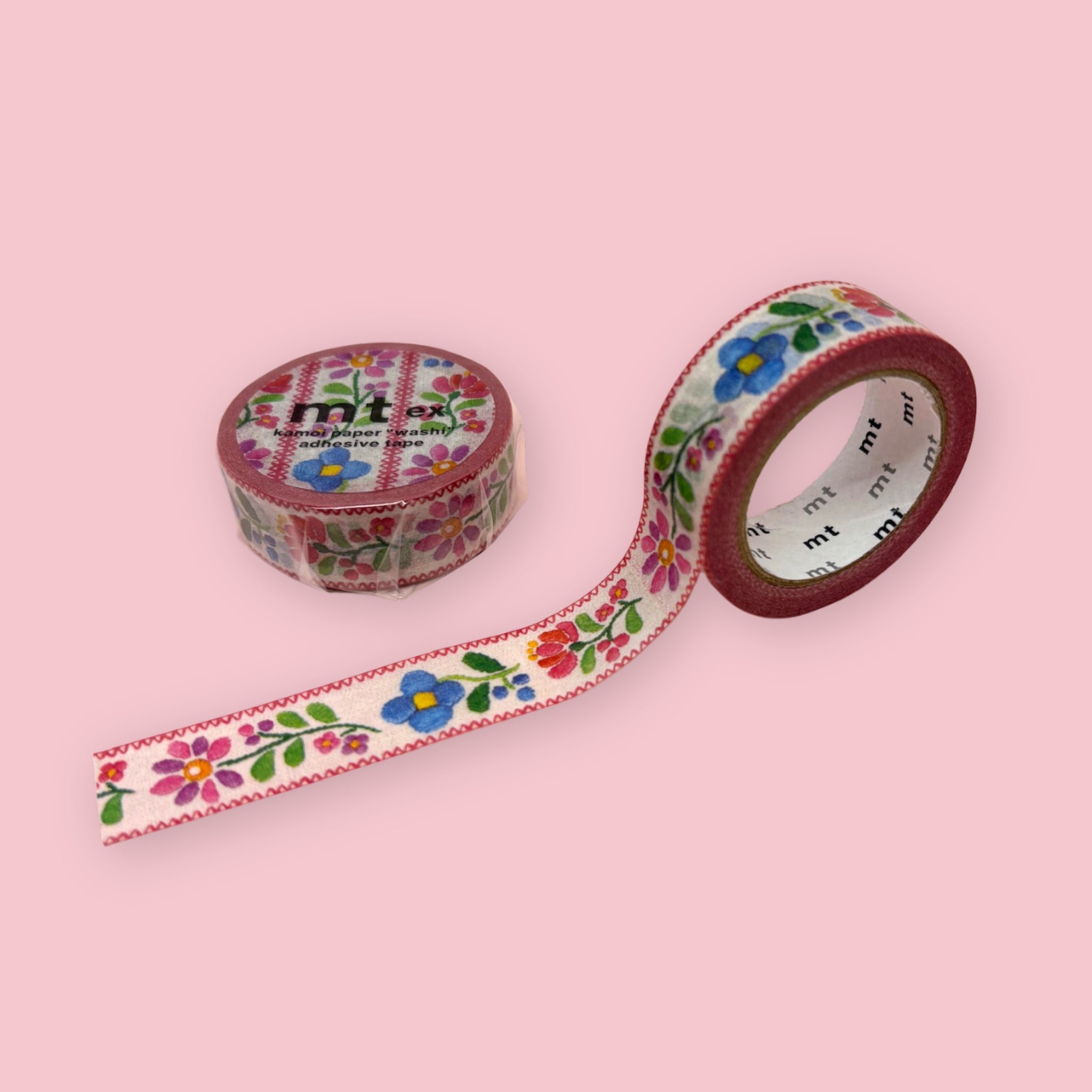 mt washi tape with a colorful floral border pattern on white, shown partly unspooled beside a wrapped roll on a pink background.