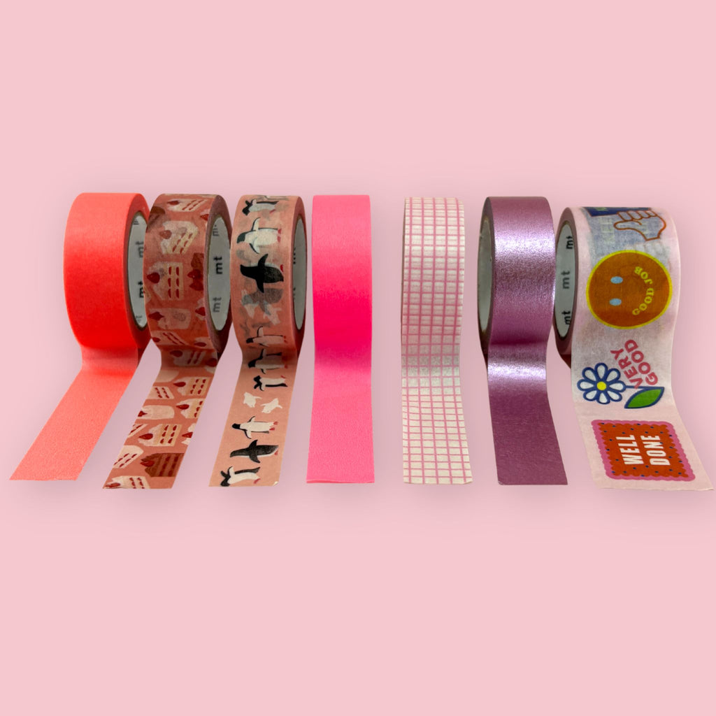 
Seven rolls of washi tape in pink tones displayed on a pink background, including solid coral, hot pink, metallic purple, grid pattern, and decorative designs featuring cakes, penguins, and sticker motifs