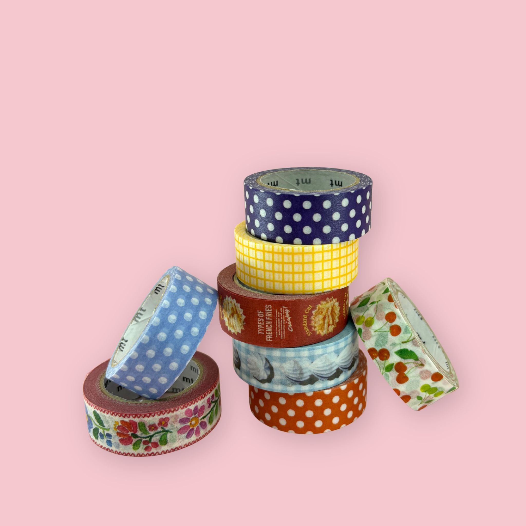 Collection of eight mt brand washi tape rolls on a pink background, featuring navy blue and white polka dots, yellow grid, rust orange polka dots, blue polka dots, light blue gingham, burgundy French fries novelty print, cherry botanical print, and an embroidered-style floral ribbon design
