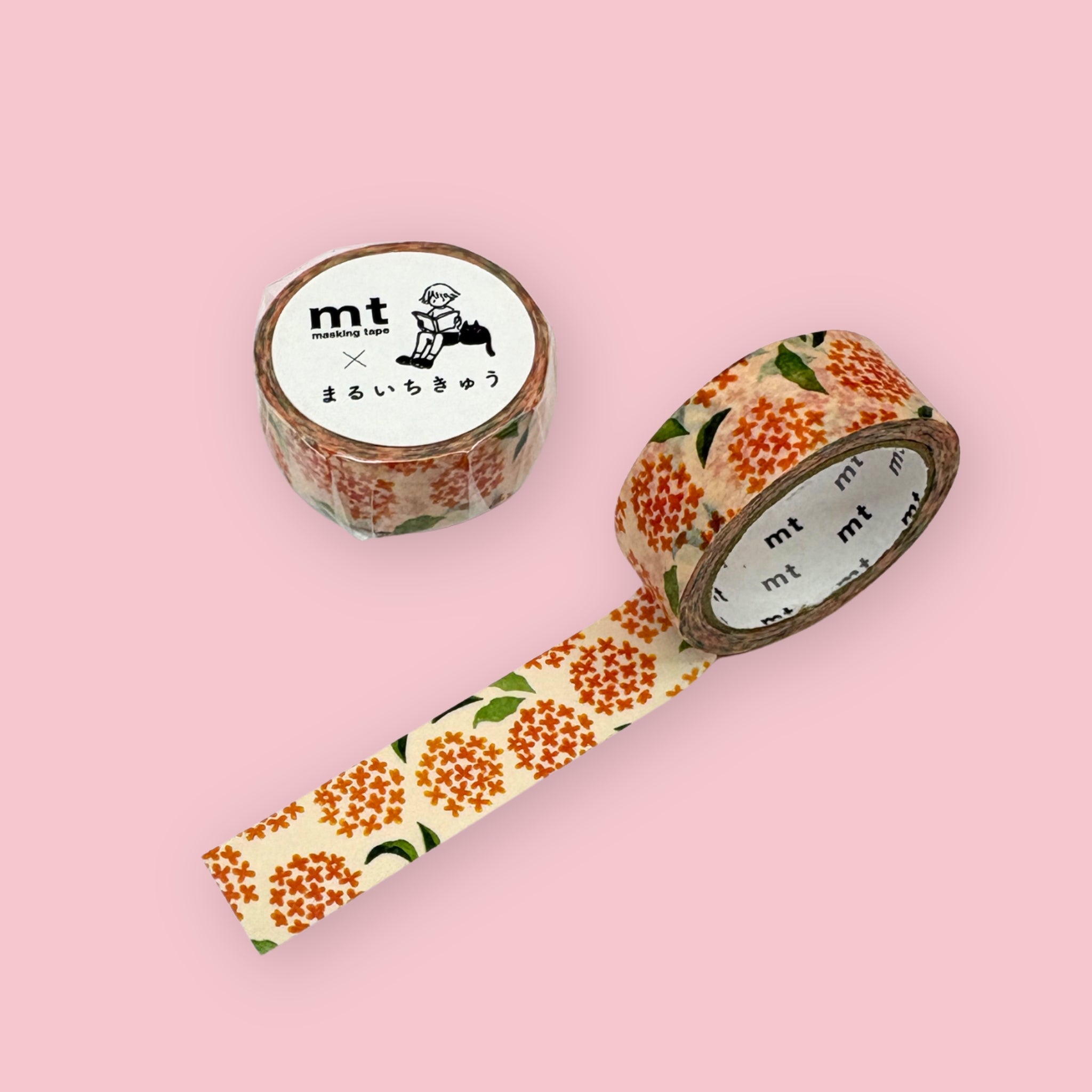 Two rolls of mt masking tape featuring clusters of small orange flowers with green leaves on a cream background, displayed on a pink surface, one roll standing upright showing the label and one partially unrolled diagonally.