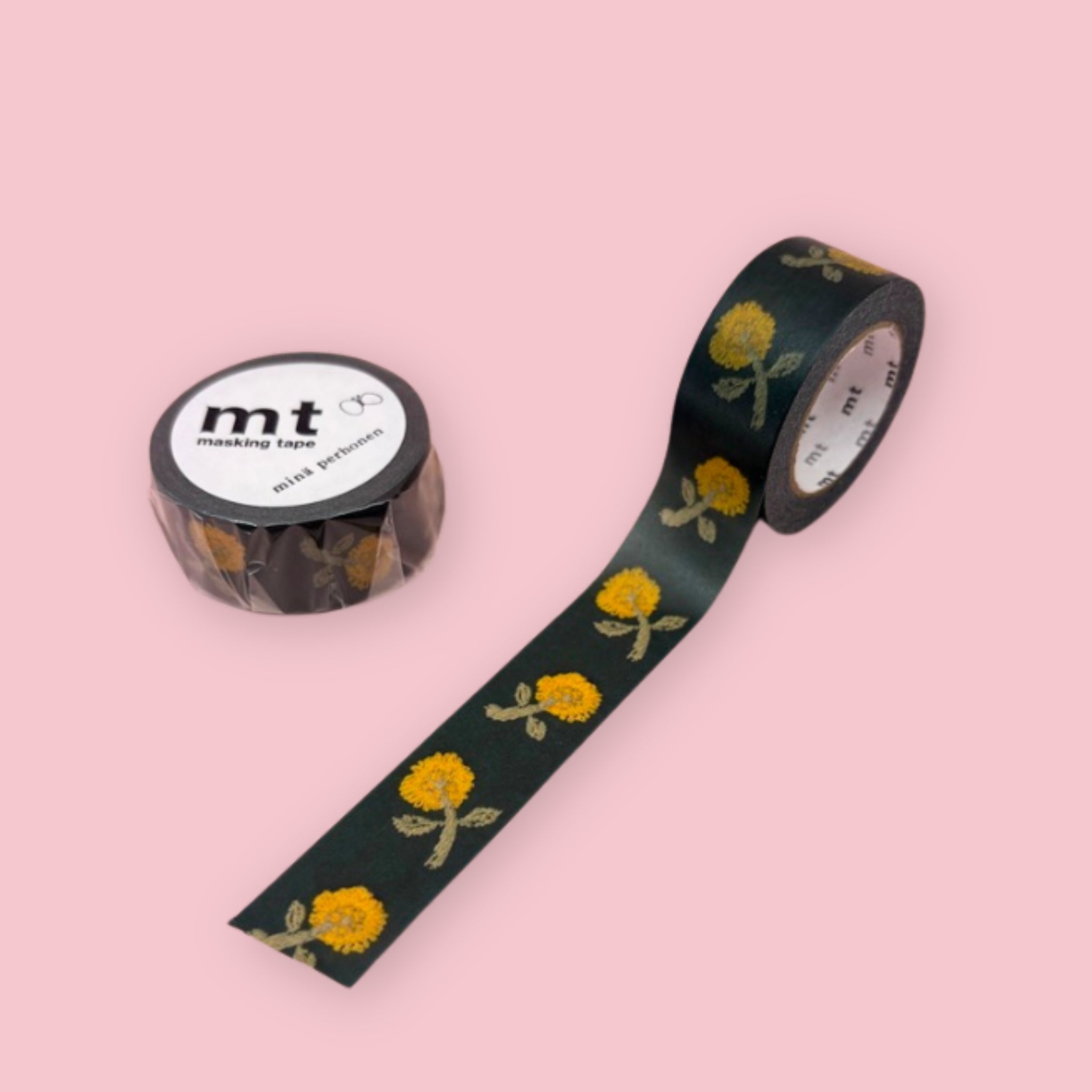 Two rolls of mt x minä perhonen masking tape featuring repeating yellow flowers on a dark green background, displayed on a pink surface — one roll standing upright showing the label, one partially unrolled diagonally.