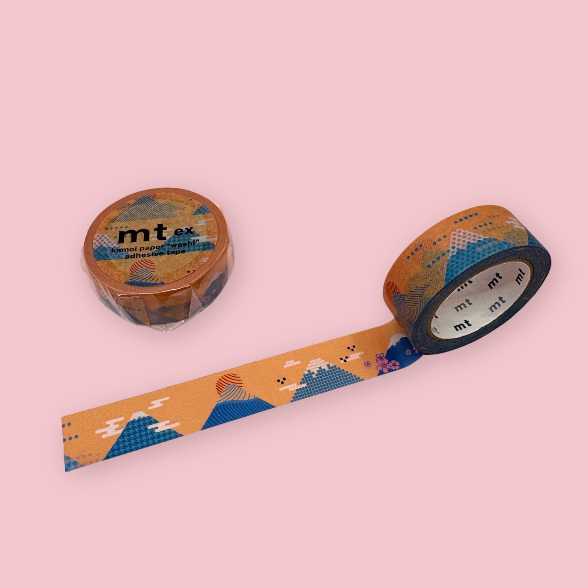 Orange mt washi tape with a blue Mt. Fuji and sunrise landscape pattern, shown partly unspooled beside a wrapped roll on a pink background.