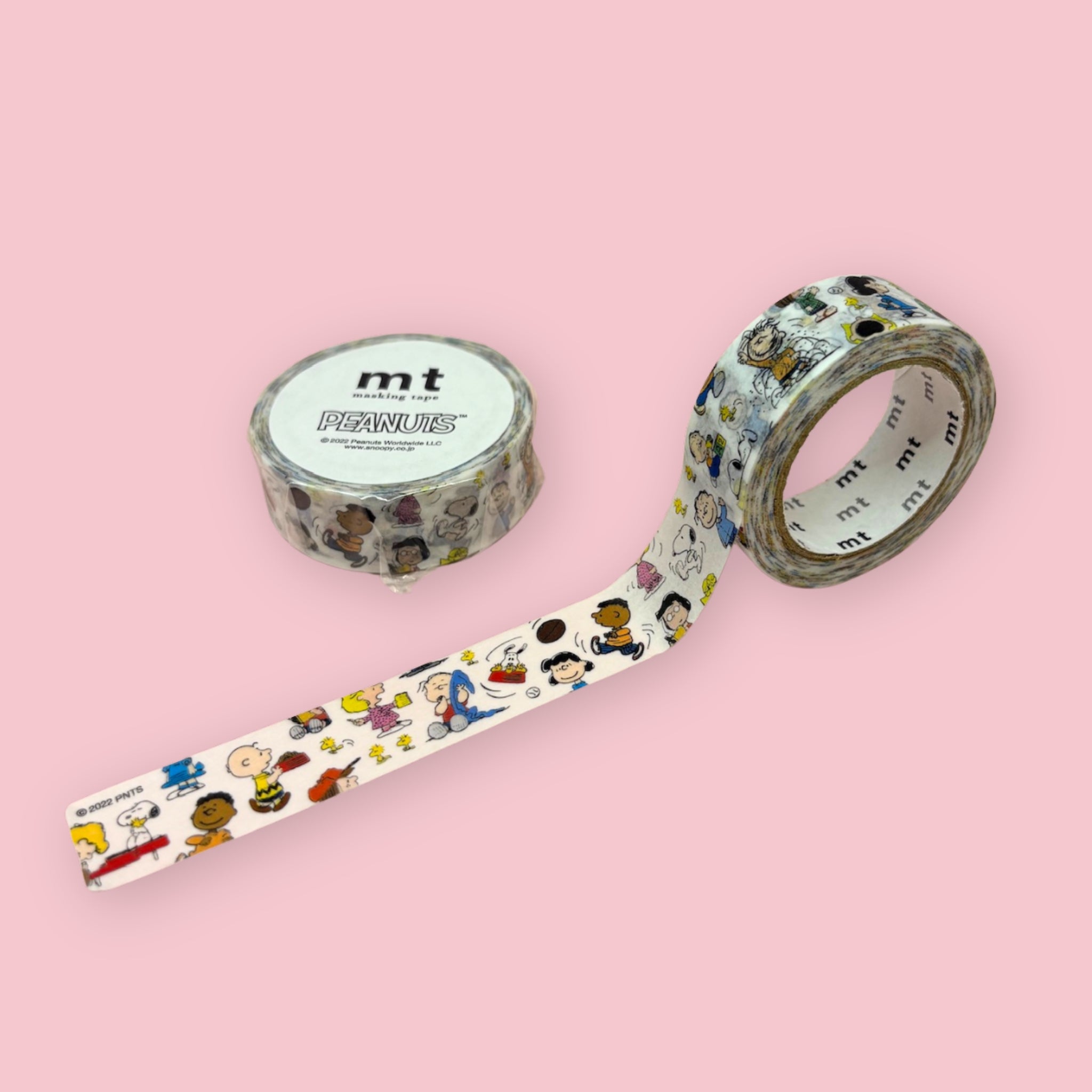 White Peanuts-themed mt washi tape with small cartoon character illustrations, shown partly unspooled beside a wrapped roll on a pink background.