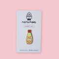 Natamori Kewpie Mayo enamel pin on its backing card with the Natamori logo, "enamel pin" label, and website handle, showing the gold-plated Kewpie mayonnaise bottle design with red lid and katakana lettering, against a pink background