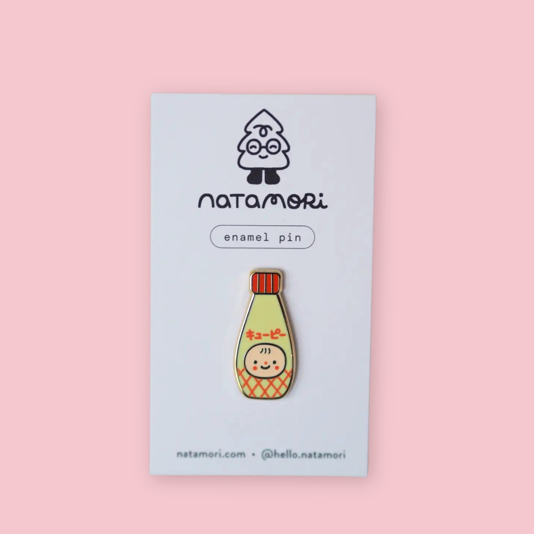 Natamori Kewpie Mayo enamel pin on its backing card with the Natamori logo, "enamel pin" label, and website handle, showing the gold-plated Kewpie mayonnaise bottle design with red lid and katakana lettering, against a pink background