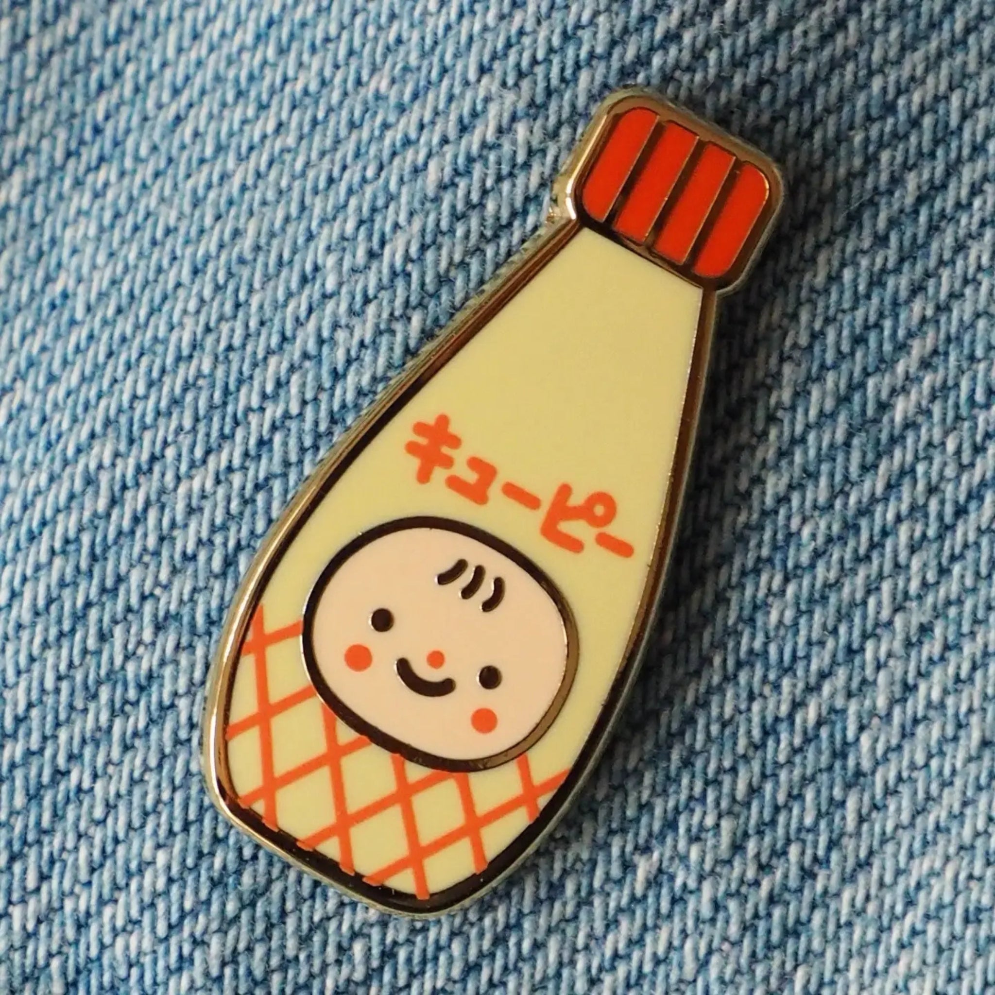 Natamori Kewpie Mayo enamel pin shaped like a Kewpie mayonnaise bottle with a red striped lid and red crosshatch pattern, pinned to a blue denim jacket