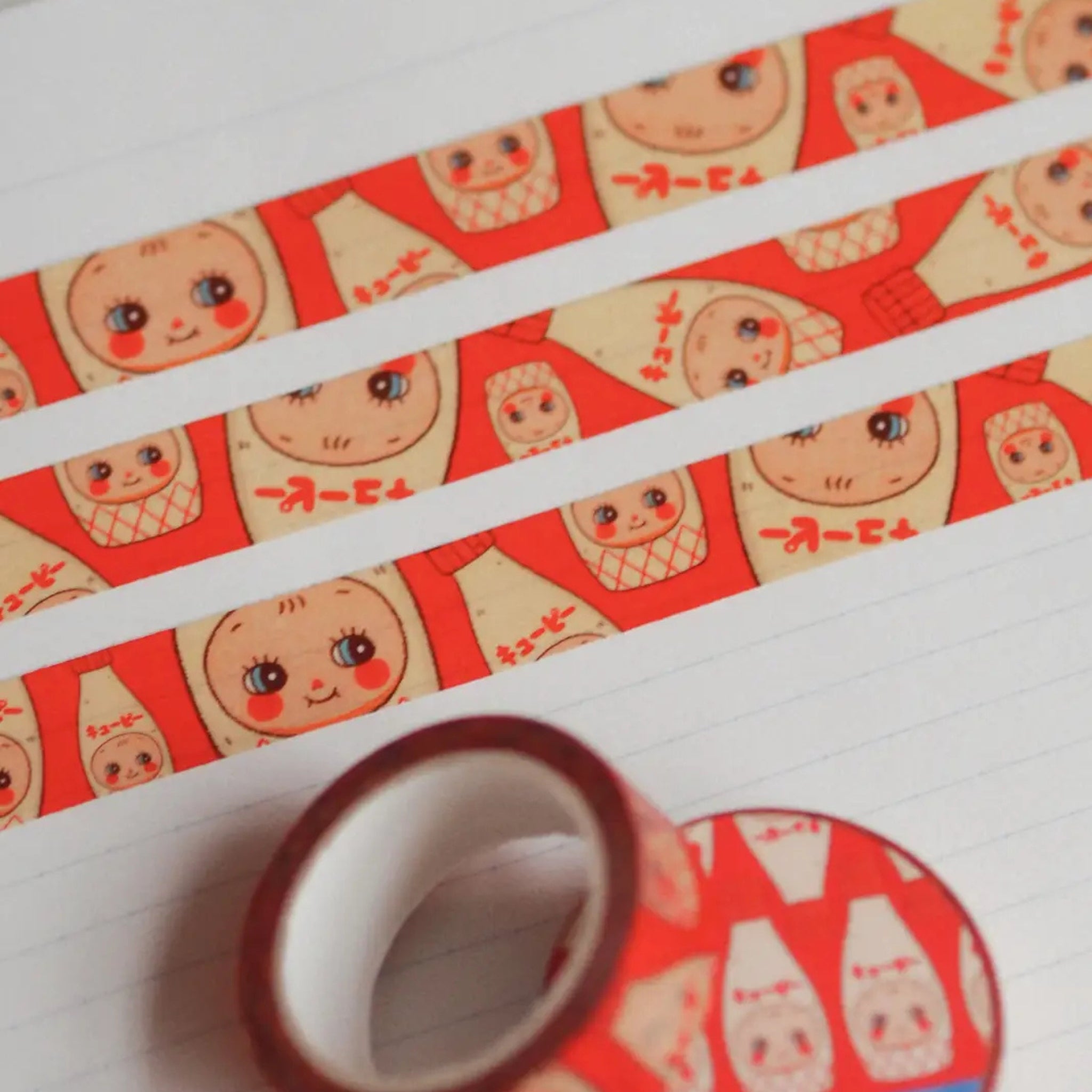 Natamori Kewpie Mayo washi tape strips printed with rows of smiling Kewpie mayonnaise bottles in red and cream, laid across a lined notebook page next to the unrolled tape roll