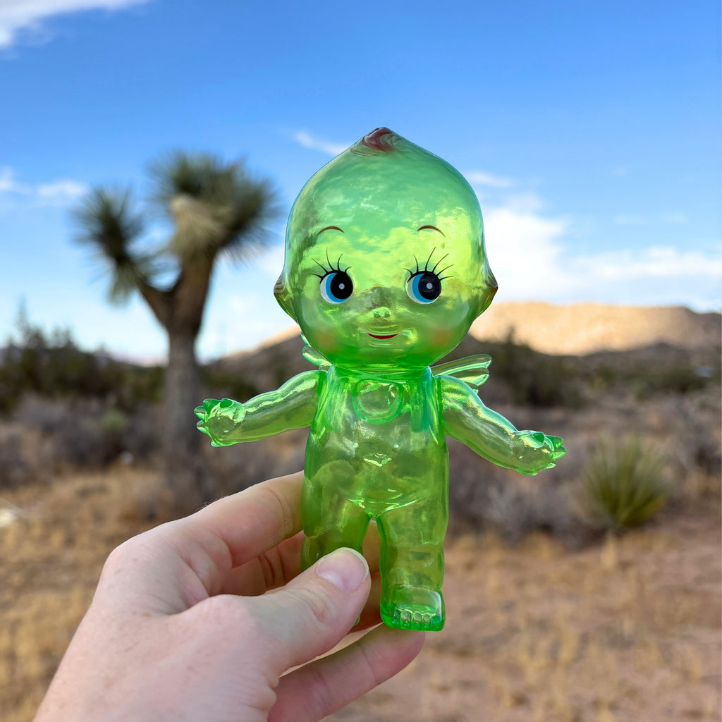 Green translucent Obitsu Kewpie doll held outdoors in the desert, with Joshua trees and mountains in the background.