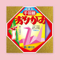Toyo Senbazuru origami paper package with a folded crane illustration in the center, gold textured borders, and Japanese text reading "Senbazuru Origami," against a pink background