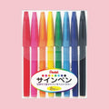 Set of eight colorful Pentel Sign Pen markers in plastic packaging, shown on a pink background.