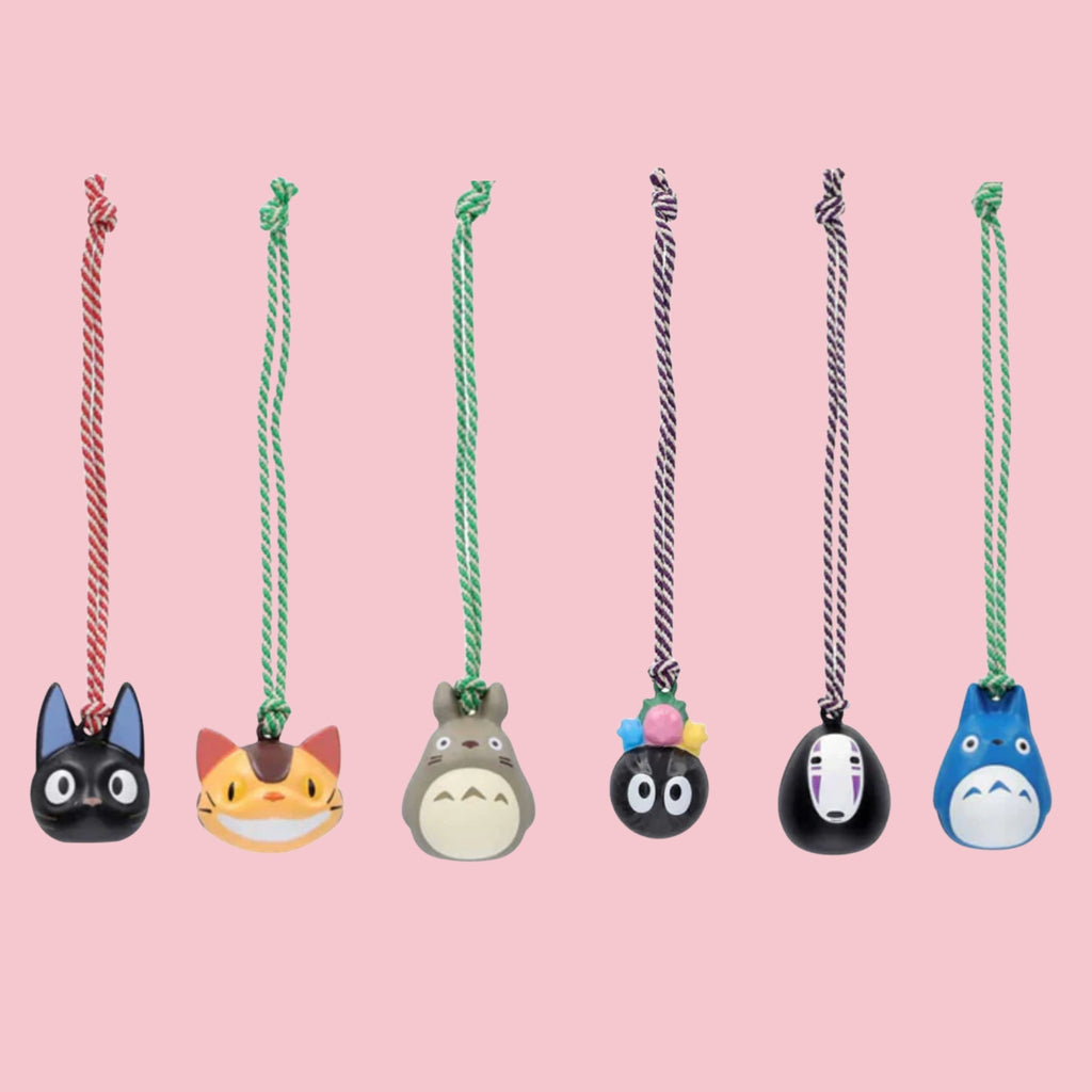 Six Studio Ghibli bell charms hanging from colored cords against a pink background, featuring Jiji, Catbus, Totoro, Soot Sprites, No-Face, and a blue Totoro variant, each with a small braided cord.