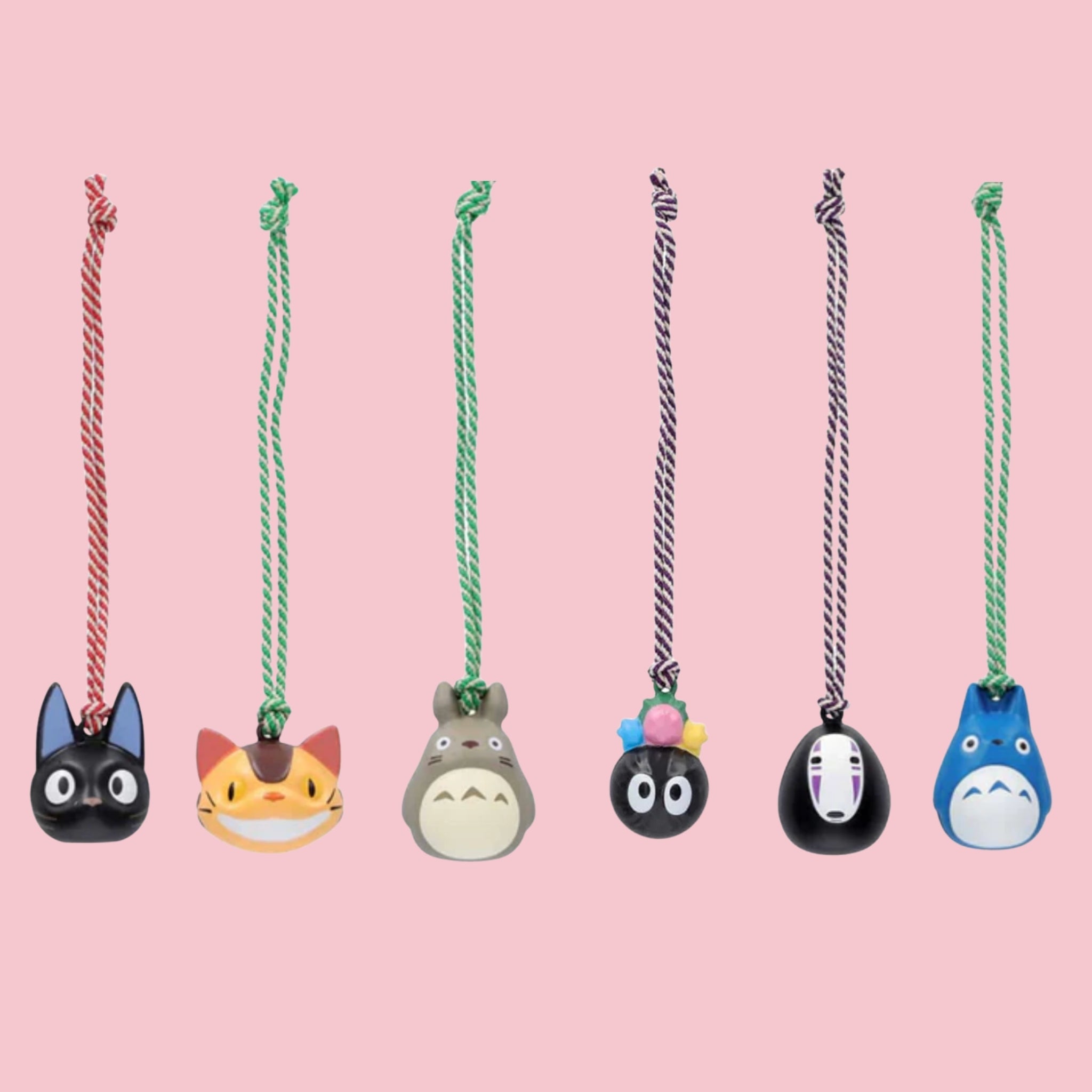 Six Studio Ghibli bell charms hanging from colored cords against a pink background, featuring Jiji, Catbus, Totoro, Soot Sprites, No-Face, and a blue Totoro variant, each with a small braided cord.