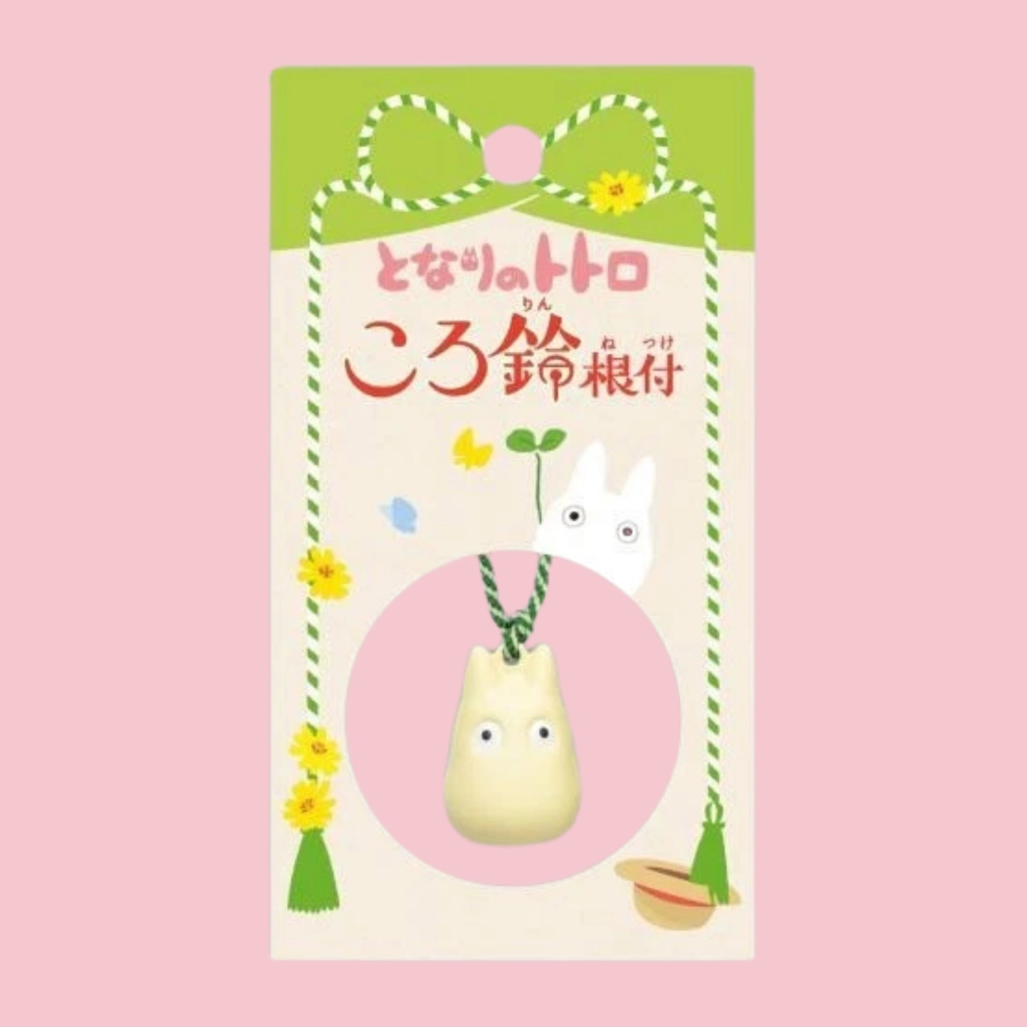 Original Japanese packaging card for the My Neighbor Totoro bell charm, featuring the character illustration and Japanese title in red text on a green and cream backdrop, with the small white Totoro bell charm shown hanging on its braided cord inside a circular pink window at the bottom of the card.